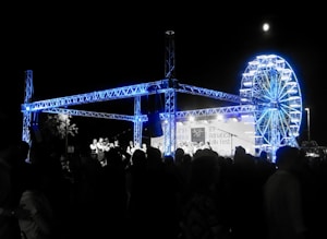 A night-time festival scene with a large crowd of people gathered around a stage. The stage features a steel truss structure illuminated in blue light. In the background, there is a brightly lit Ferris wheel. A banner on the stage reads Adriatica Folk Fest.