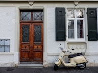 A cream-colored Vespa scooter is parked in front of a building. The building features a large, ornate wooden double door with intricate metalwork on the left side. To the right of the door is a window with white frames and dark green shutters on either side, one of which is closed. The exterior walls of the building are textured and light-colored. Mounted on the left side of the image are several mailboxes.