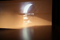 A soft, ethereal light cascades through an archway, illuminating the phrase 'ALMIGHTY GOD' projected in the center. The surroundings are warm and hazy, creating a serene and contemplative atmosphere.