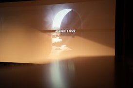 A soft, ethereal light cascades through an archway, illuminating the phrase 'ALMIGHTY GOD' projected in the center. The surroundings are warm and hazy, creating a serene and contemplative atmosphere.