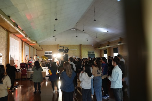 A large group of people are gathered in a hall with a high ceiling and brick walls. To the left, a band is playing with musicians using instruments such as keyboards and guitars. The lighting is focused on the band, while attendees stand and observe, some with their hands raised.