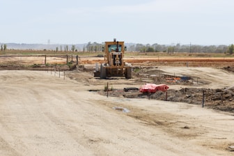 A construction site with a large vehicle, resembling a bulldozer or grader, positioned in the center. The landscape is expansive with dirt paths and scattered construction materials. The background features open fields and a line of trees stretching across the horizon.