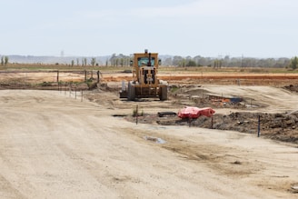 A construction site with a large vehicle, resembling a bulldozer or grader, positioned in the center. The landscape is expansive with dirt paths and scattered construction materials. The background features open fields and a line of trees stretching across the horizon.