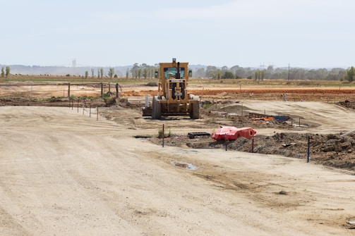 A construction site with a large vehicle, resembling a bulldozer or grader, positioned in the center. The landscape is expansive with dirt paths and scattered construction materials. The background features open fields and a line of trees stretching across the horizon.