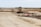 A construction site with a large vehicle, resembling a bulldozer or grader, positioned in the center. The landscape is expansive with dirt paths and scattered construction materials. The background features open fields and a line of trees stretching across the horizon.