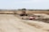 A construction site with a large vehicle, resembling a bulldozer or grader, positioned in the center. The landscape is expansive with dirt paths and scattered construction materials. The background features open fields and a line of trees stretching across the horizon.