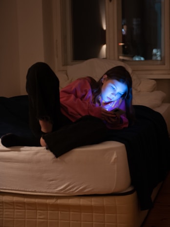 A person is lying on a bed in a dimly lit room, intensely focused on a smartphone. The glow from the phone illuminates their face. The environment appears calm and cozy, with a window in the background showing a hint of outside light.