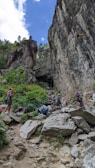 A group of people are engaged in rock climbing on a large cliff face in a natural setting. Several individuals are climbing the rock wall, while others are gathered at its base, equipped with climbing gear. The landscape is rugged, with large rocks and green vegetation surrounding the area. The sky is partially cloudy, contributing to the outdoor adventure ambiance.