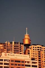 A cityscape featuring the prominent Westfield Tower, surrounded by modern high-rise buildings. The Bank of China building is visible in the foreground with its sign in both Chinese and English. The scene is set against a clear evening sky, giving the buildings a warm, golden glow.