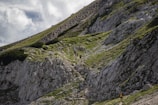 A group of hikers ascends a steep, rocky mountain path covered with patches of green grass and small shrubs. The mountainside is rugged and the sky is partly cloudy, adding a dramatic effect to the landscape.