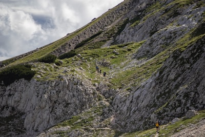 A group of hikers ascends a steep, rocky mountain path covered with patches of green grass and small shrubs. The mountainside is rugged and the sky is partly cloudy, adding a dramatic effect to the landscape.