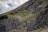 A group of hikers ascends a steep, rocky mountain path covered with patches of green grass and small shrubs. The mountainside is rugged and the sky is partly cloudy, adding a dramatic effect to the landscape.