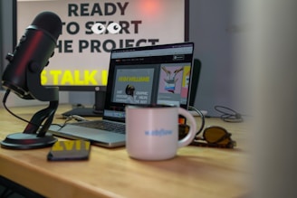 A microphone is positioned near a laptop on a wooden desk, with sunglasses and a white mug displaying the word 'Webflow'. The background features a computer monitor displaying partially obscured text, suggesting a workspace environment.