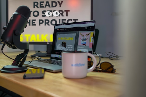 A microphone is positioned near a laptop on a wooden desk, with sunglasses and a white mug displaying the word 'Webflow'. The background features a computer monitor displaying partially obscured text, suggesting a workspace environment.