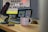 A microphone is positioned near a laptop on a wooden desk, with sunglasses and a white mug displaying the word 'Webflow'. The background features a computer monitor displaying partially obscured text, suggesting a workspace environment.