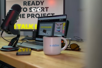 A microphone is positioned near a laptop on a wooden desk, with sunglasses and a white mug displaying the word 'Webflow'. The background features a computer monitor displaying partially obscured text, suggesting a workspace environment.