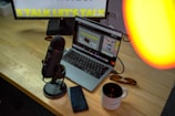 A workspace is set with a laptop displaying a graphic design interface. A microphone is positioned on a wooden desk beside a smartphone and a coffee mug. A monitor in the background partially shows text, possibly for a presentation. There is a warm light source illuminating part of the scene.