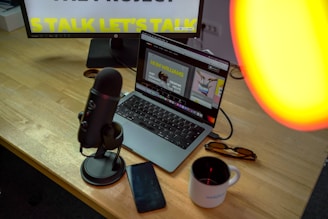A workspace is set with a laptop displaying a graphic design interface. A microphone is positioned on a wooden desk beside a smartphone and a coffee mug. A monitor in the background partially shows text, possibly for a presentation. There is a warm light source illuminating part of the scene.