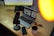 A workspace is set with a laptop displaying a graphic design interface. A microphone is positioned on a wooden desk beside a smartphone and a coffee mug. A monitor in the background partially shows text, possibly for a presentation. There is a warm light source illuminating part of the scene.