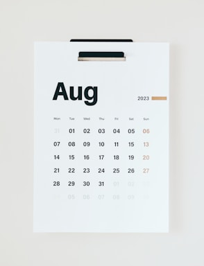 A minimalist wall calendar featuring the month of August 2023. The calendar displays the days of the week starting from Monday and highlights Sundays in a different color. It is suspended on a white wall using a black clip.