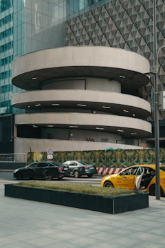 A modern urban scene featuring a multi-level parking garage with an architectural facade of geometric patterns. Several cars, including a yellow taxi, are visible on the road next to a sidewalk with patches of grass and a 'no parking' sign.