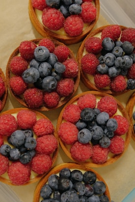 A collection of small fruit tarts topped generously with fresh raspberries and blueberries, arranged neatly on a beige surface.