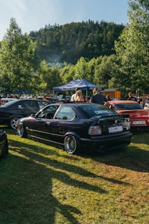 Several cars are parked in an open grassy area near a forested hillside. A dark-colored car is prominently displayed in the foreground, with people gathered around, possibly at an outdoor car event. A blue tent canopy with branding is visible among the crowd.