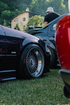 A close-up view of two parked cars on grass, featuring distinct and polished rims. A person wearing a light-colored hat is standing next to one of the cars. In the background, there is a yellow house with a curved roof surrounded by green trees.
