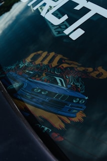 A car dashboard with an artistic design featuring a blue sports car graphic. The image includes vibrant colors and dynamic elements like flames and bold text. The design is partially reflected on a surface, possibly on a car window.