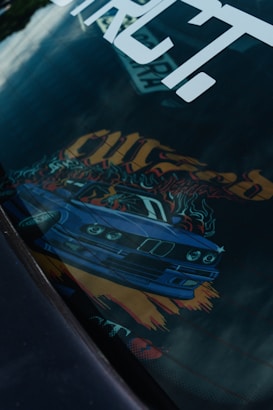 A car dashboard with an artistic design featuring a blue sports car graphic. The image includes vibrant colors and dynamic elements like flames and bold text. The design is partially reflected on a surface, possibly on a car window.