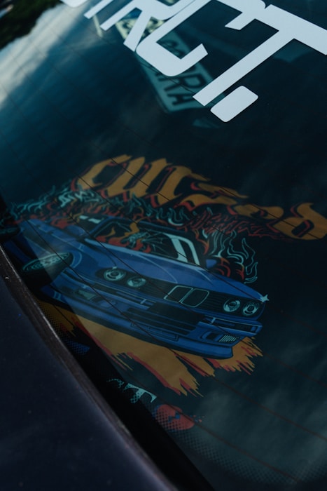 A car dashboard with an artistic design featuring a blue sports car graphic. The image includes vibrant colors and dynamic elements like flames and bold text. The design is partially reflected on a surface, possibly on a car window.