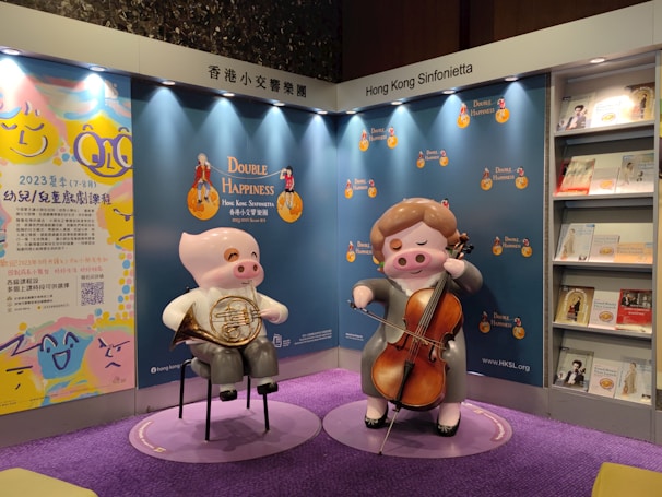 The image features a display booth for the Hong Kong Sinfonietta. Two cartoonish pig statues are prominently shown playing musical instruments, one holding a French horn and the other a cello. The booth backdrop showcases the 'Double Happiness' theme with light blue and orange hues. There are brochures or booklets arranged on shelves to the right, and colorful artwork with text on the left side. The floor is covered with a vibrant purple carpet.