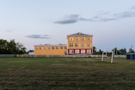 A three-story yellow building with red accents is situated next to an open grassy field. A set of goalposts without a net is visible in the foreground, and trees line the background. The sky is clear with a few scattered clouds.