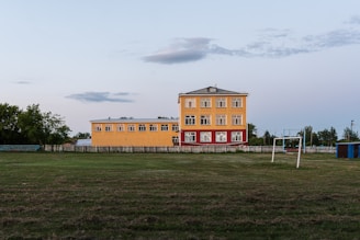 A three-story yellow building with red accents is situated next to an open grassy field. A set of goalposts without a net is visible in the foreground, and trees line the background. The sky is clear with a few scattered clouds.