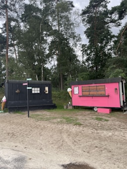 Two small trailers are situated on a sandy area surrounded by tall, green trees. One trailer is painted black with a window and some decorations, while the other is bright pink and appears to be a food stall, with an ice cream cone sign and a menu board.