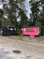 Two small trailers are situated on a grassy area with trees in the background. One trailer is painted black and has a rustic look with decorative arrangements on it. The other is bright pink with a service window, suggesting it might be used for business purposes, possibly serving food or beverages. Between the trailers, a small wooden signpost is visible.