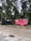 Two small trailers are situated on a grassy area with trees in the background. One trailer is painted black and has a rustic look with decorative arrangements on it. The other is bright pink with a service window, suggesting it might be used for business purposes, possibly serving food or beverages. Between the trailers, a small wooden signpost is visible.