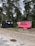 Two small trailers are situated on a grassy area with trees in the background. One trailer is painted black and has a rustic look with decorative arrangements on it. The other is bright pink with a service window, suggesting it might be used for business purposes, possibly serving food or beverages. Between the trailers, a small wooden signpost is visible.