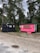 Two small trailers are situated on a grassy area with trees in the background. One trailer is painted black and has a rustic look with decorative arrangements on it. The other is bright pink with a service window, suggesting it might be used for business purposes, possibly serving food or beverages. Between the trailers, a small wooden signpost is visible.