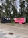 Two small trailers are situated on a grassy area with trees in the background. One trailer is painted black and has a rustic look with decorative arrangements on it. The other is bright pink with a service window, suggesting it might be used for business purposes, possibly serving food or beverages. Between the trailers, a small wooden signpost is visible.