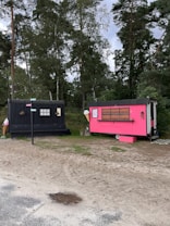 Two small trailers are situated on a grassy area with trees in the background. One trailer is painted black and has a rustic look with decorative arrangements on it. The other is bright pink with a service window, suggesting it might be used for business purposes, possibly serving food or beverages. Between the trailers, a small wooden signpost is visible.