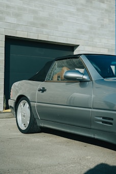 A silver convertible car is parked near a large, industrial-style building with a textured, brick-like facade. The car's top is down, showing the interior and the reflections on its metallic surface.