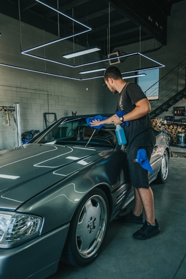 A person is cleaning a sleek, silver car with a blue cloth and spray bottle inside a well-lit garage. The garage features modern, geometric ceiling lights and has a partially visible staircase in the background.