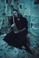A person sitting on a floor covered with newspapers, holding a string instrument while appearing to be asleep or in deep thought. The walls are also adorned with newspaper clippings, creating an enclosed, intimate space.