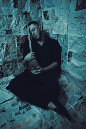 A person sitting on a floor covered with newspapers, holding a string instrument while appearing to be asleep or in deep thought. The walls are also adorned with newspaper clippings, creating an enclosed, intimate space.