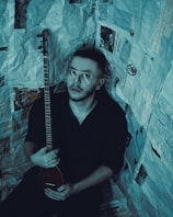 A person with short hair and glasses holds a stringed musical instrument. The individual is surrounded by walls covered in newspaper pages, creating an enclosed, creative atmosphere.