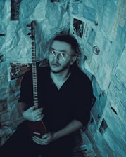 A person with short hair and glasses holds a stringed musical instrument. The individual is surrounded by walls covered in newspaper pages, creating an enclosed, creative atmosphere.