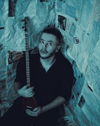 A person with short hair and glasses holds a stringed musical instrument. The individual is surrounded by walls covered in newspaper pages, creating an enclosed, creative atmosphere.
