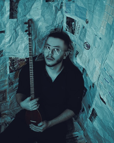 A person with short hair and glasses holds a stringed musical instrument. The individual is surrounded by walls covered in newspaper pages, creating an enclosed, creative atmosphere.