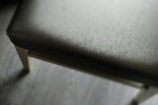 A close-up view of a padded chair seat with a texture similar to fabric. The seat is in focus while the background is blurred. The chair legs are wooden, and the floor beneath is made of light-colored wood.